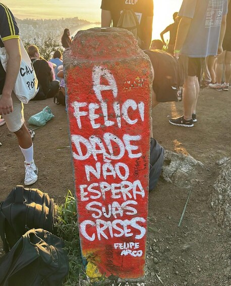 A red sign with white graffiti saying 'A FELICI DADE NAO ESPERA SUAS CRISES' by Felipe Arco. 3