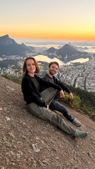 Two people sitting on a rocky outcrop overlooking a cityscape with mountains and a river. 3