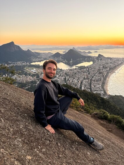 A person sitting on a rocky outcrop overlooking a cityscape with a river and mountains in the background. 1