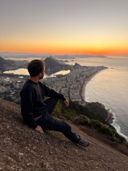 A person sitting on a rocky outcrop overlooking a coastal cityscape during sunset. 2