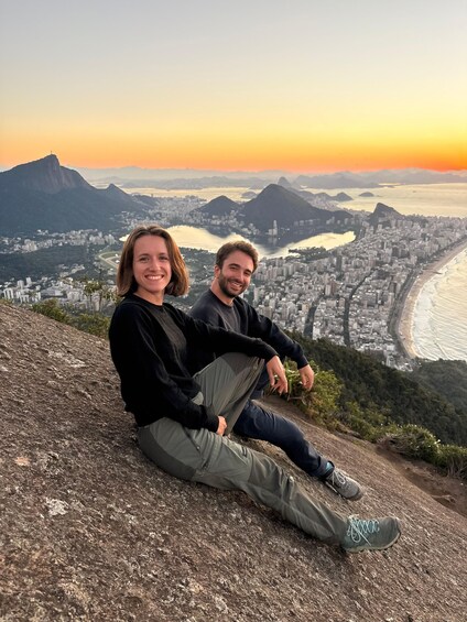 Two people sitting on a rocky outcrop overlooking a cityscape with mountains and a river. 5