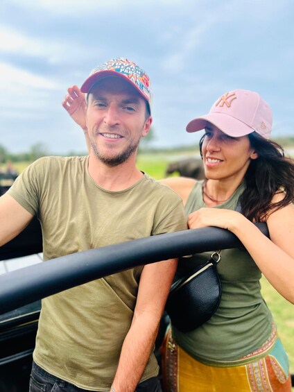Two people in a vehicle, one wearing a green t-shirt and a colorful cap, the other in a green top and a pink cap. 1