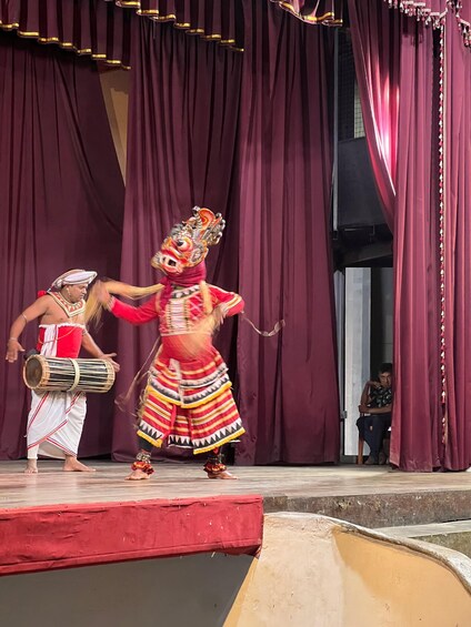A performer in a colorful costume with a headpiece, playing a drum on a stage with maroon curtains. 4