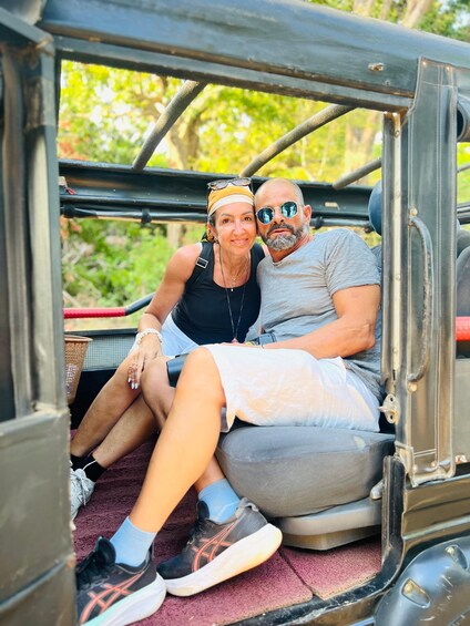 Two people sitting in a vehicle's open door, with greenery in the background. 1