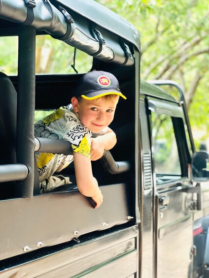 A child leaning out of a vehicle window, wearing a cap and a patterned shirt. 1