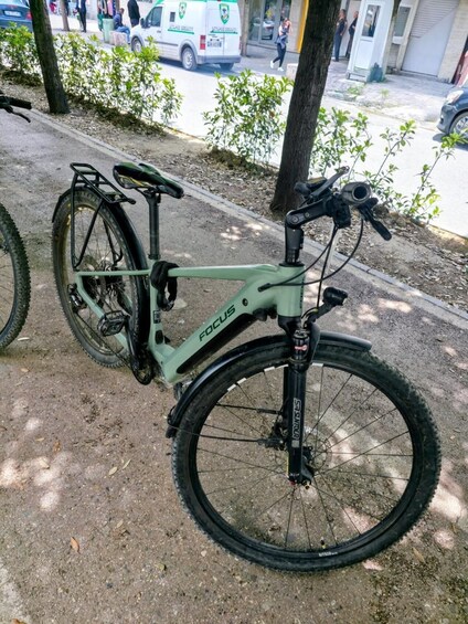 A green Focus mountain bike parked on a sidewalk with a tree and a street in the background. 3
