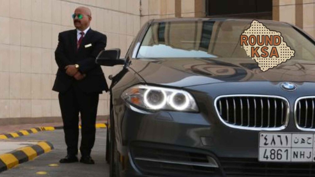 A man in a suit standing beside a black BMW car with a license plate. 3