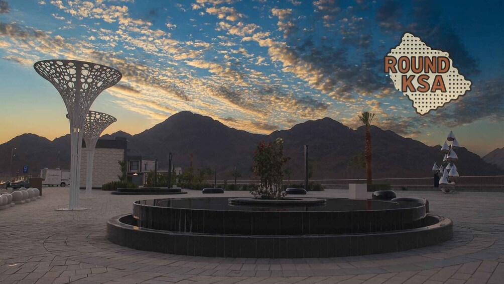A roundabout with a modern sculpture, a fountain, and mountains in the background. 4