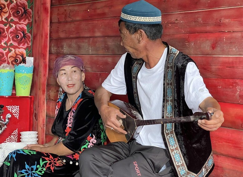 A man playing a stringed instrument, a woman seated beside him, and a red wooden structure with decorative elements. 2
