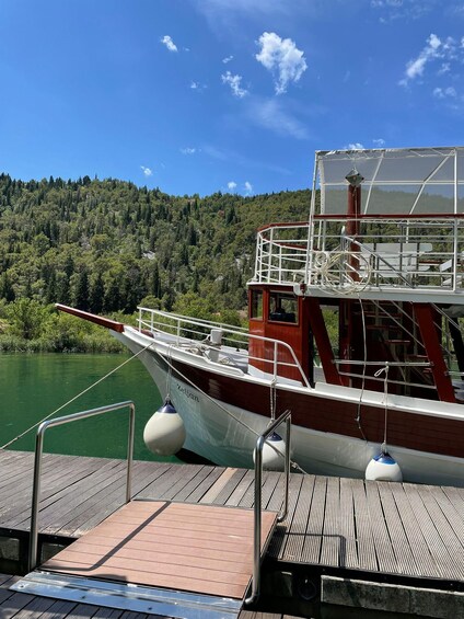 A boat docked at a pier with a forested hillside in the background. 1
