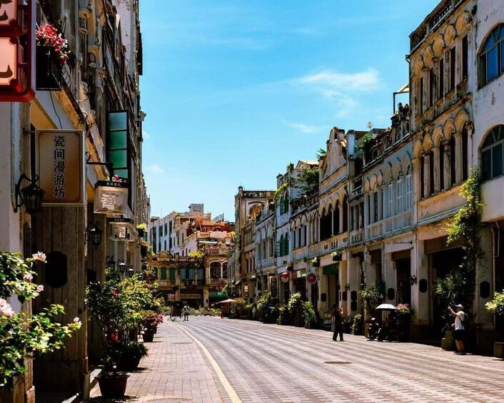 A sunlit street lined with historic buildings, potted plants, and a clear blue sky. 2