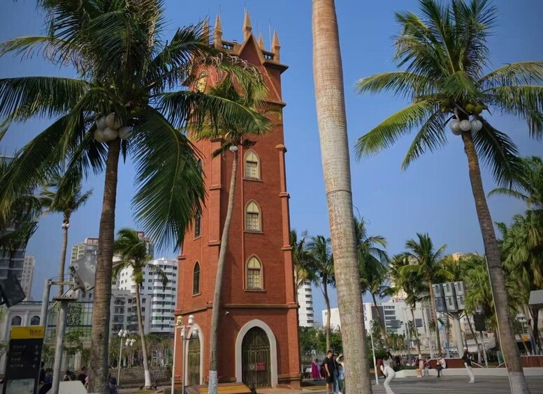 A red brick tower with arched windows and a pointed roof, surrounded by palm trees and modern buildings. 1