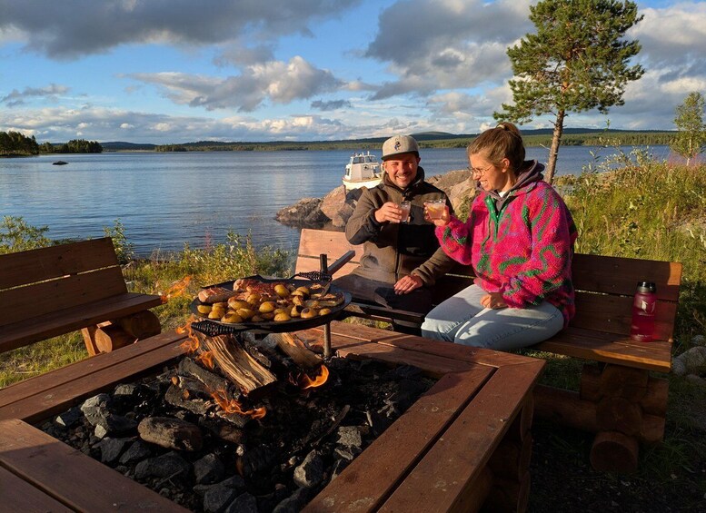 Two people sitting on a bench by a campfire with grilled food, overlooking a lake. 5