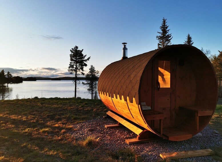 A wooden barrel-shaped cabin with a chimney, situated on a gravel area near a lake with trees in the background. 1
