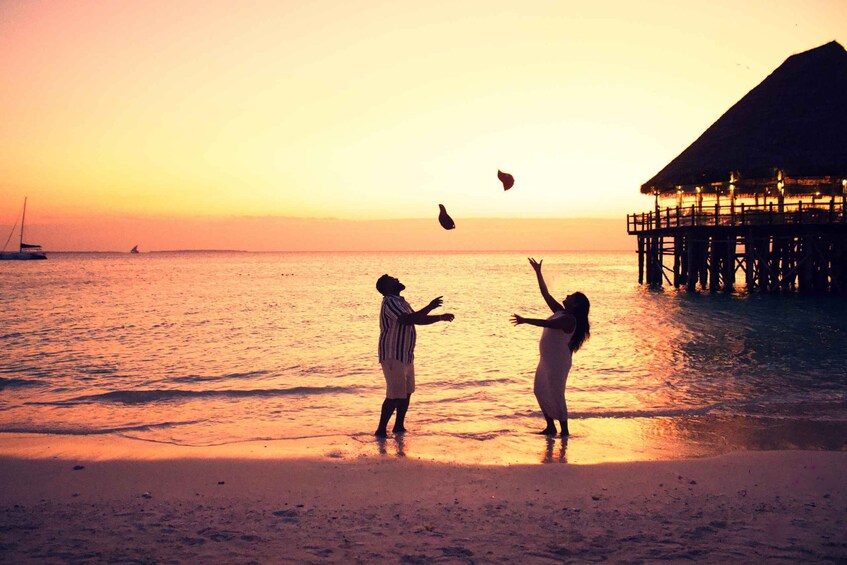 A sunset over a beach with two people playing with a kite, a boat in the distance, and a thatched-roof structure on stilts. 5