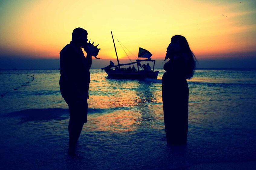 Silhouettes of two people standing on a beach with a boat in the background during sunset. 2