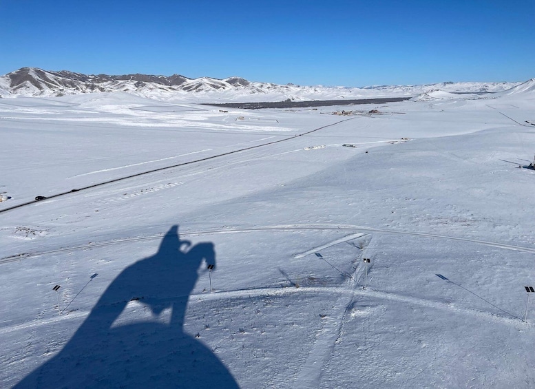 A snowy landscape with a shadow of a person on the ground, a road, and distant mountains. 5