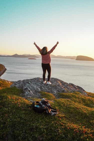 A person standing on a rocky outcrop with arms raised, overlooking a calm sea with a ship in the distance. 4