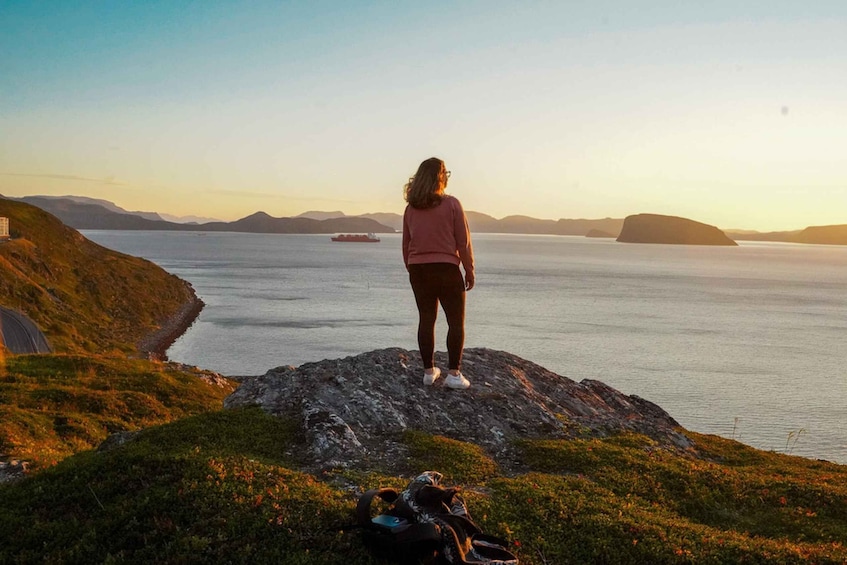 A person standing on a rocky outcrop overlooking a calm sea with a ship in the distance and hills in the background. 3