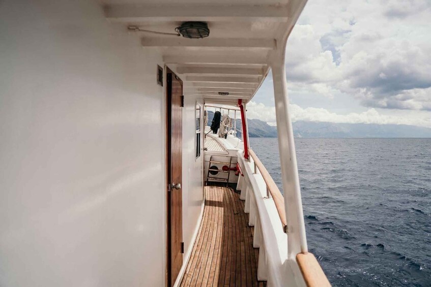 A ship's deck with a wooden walkway, white railings, and a view of the sea and distant mountains. 1