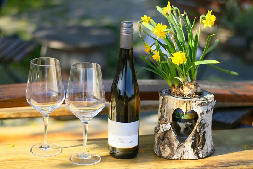 Two empty wine glasses, a bottle of 2019 Chardonnay Blanc, and a daffodil arrangement in a heart-shaped planter on a wooden table. 1