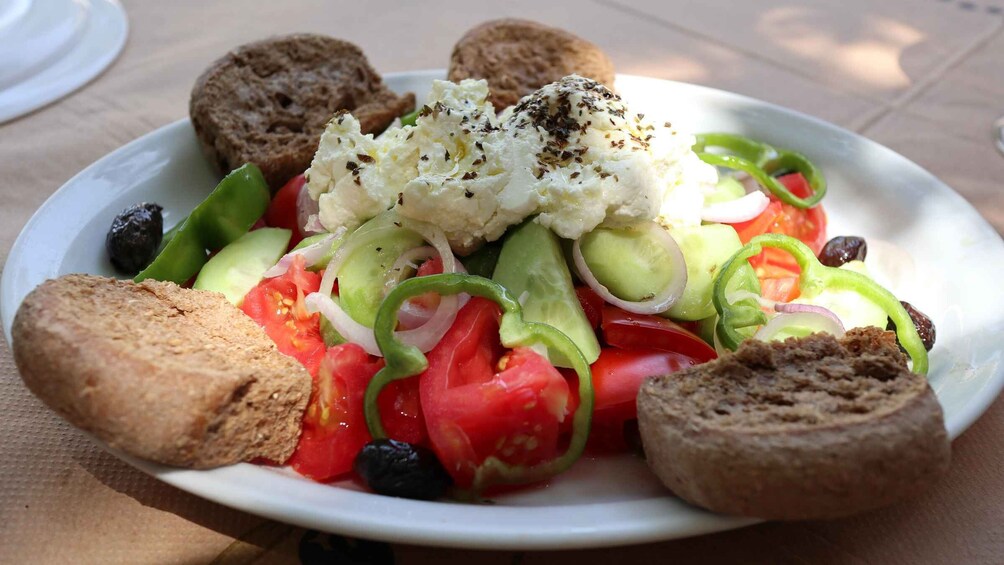 A plate of salad with tomatoes, cucumbers, peppers, and olives, topped with a white creamy substance and herbs, accompanied by slices of bread. 2
