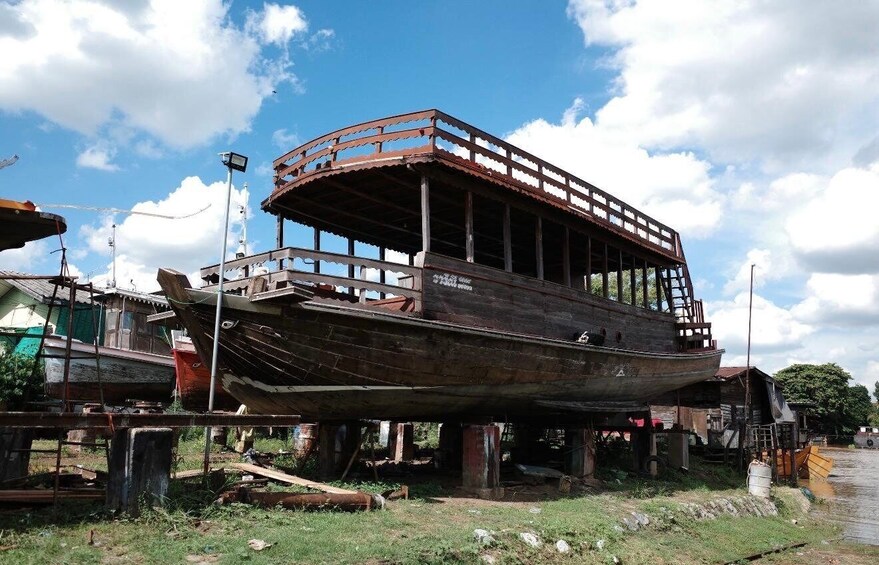 A large, rusted boat with a wooden superstructure, partially elevated on supports, in a rural setting with other boats and structures in the background. 3