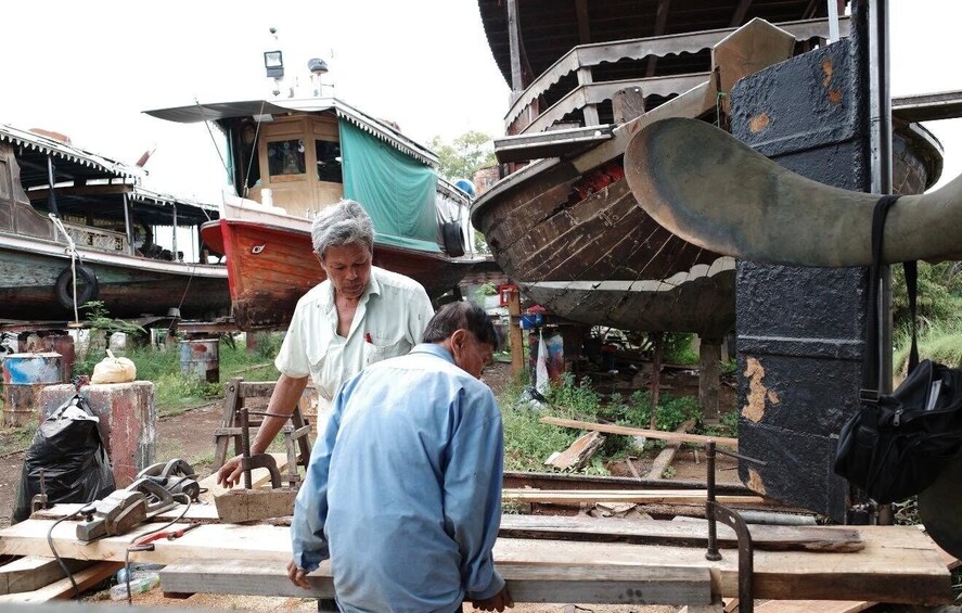 Two men working with wooden planks and tools in a workshop with boats and a large metal structure in the background. 5