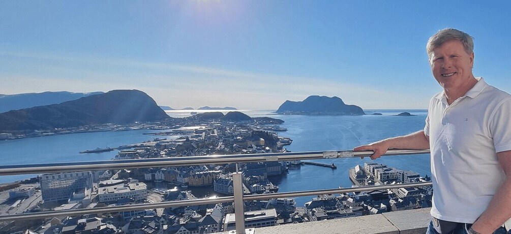 A man standing on a balcony overlooking a coastal city with a bridge and mountains in the background. 1