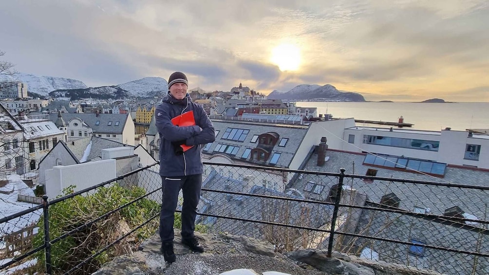 A person standing on a rocky ledge overlooking a coastal town with snow-covered mountains in the background. 4