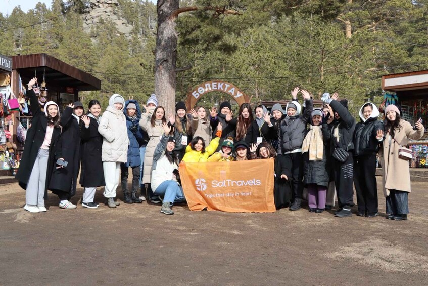 A group of people posing in front of a sign with Cyrillic text, with a banner reading 'SatTravels' and a tree in the background. 3