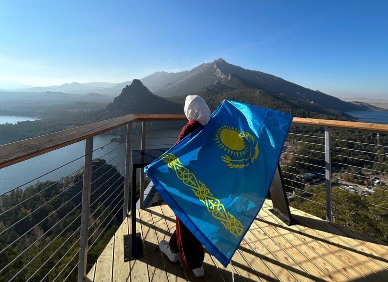 A person holding a blue flag with yellow designs, overlooking a mountainous landscape with a body of water below. 4