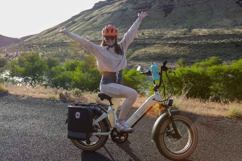 A person on a white electric bike with a backpack, arms raised, on a road with greenery and hills in the background. 5