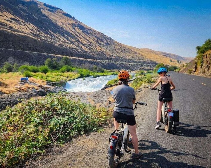 Two people on bicycles near a river with mountains in the background. 2