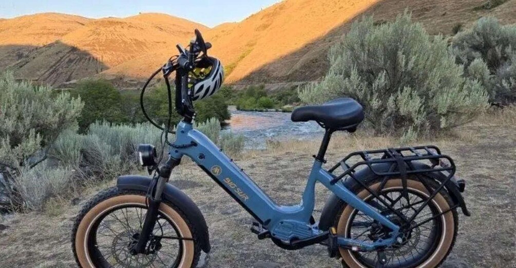 A blue electric bike with a front basket and a rear rack, parked by a river with mountains in the background. 3