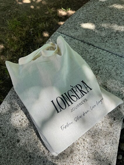 A white tote bag with 'LONG'SERA JOURNEYS' and 'Explore Slow Down, Live Longer' printed on it, placed on a stone bench. 3