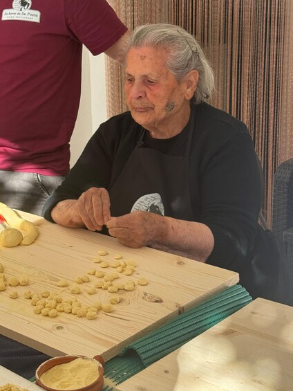 An elderly person seated at a wooden table, working with dough. 1