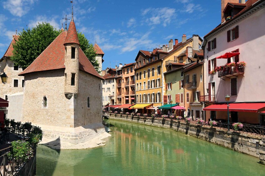 A historic tower with a conical roof, a canal with a stone wall, and colorful buildings with balconies and flower boxes. 1