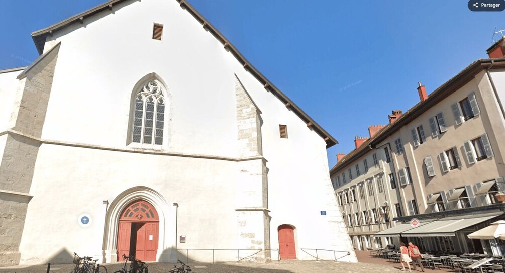 A white church with a red door and a large window, adjacent to a row of buildings with outdoor seating. 5