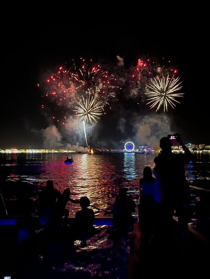 Fireworks over water with reflections, a Ferris wheel, and silhouettes of people. 1