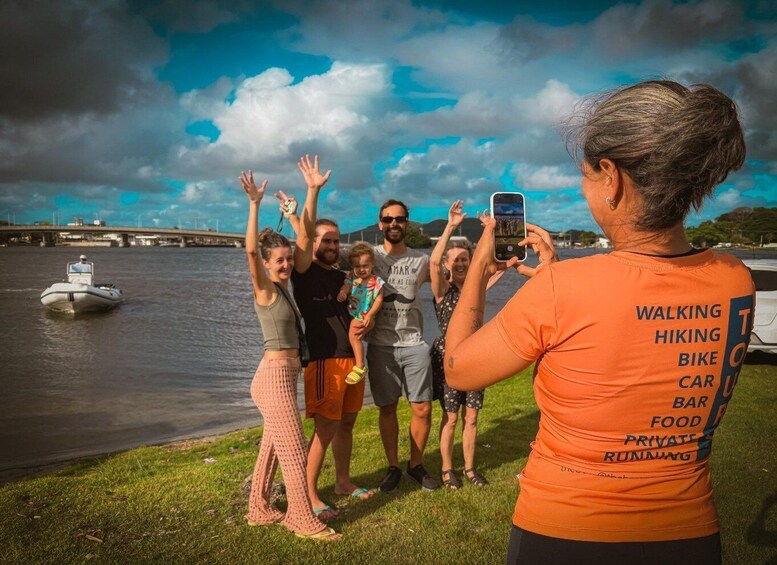 A group of people by a river with a boat in the water, a bridge in the background, and a person taking a photo. 5