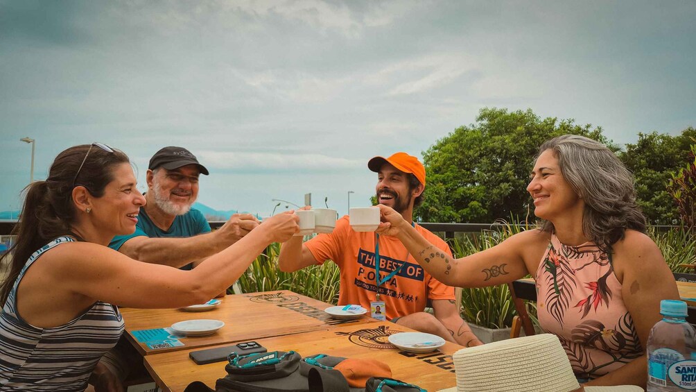 Four people toasting with cups at a wooden table outdoors with greenery in the background. 2