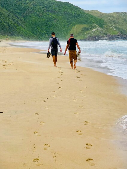 Two people walking on a sandy beach with footprints, a green hillside in the background, and waves crashing onto the shore. 3