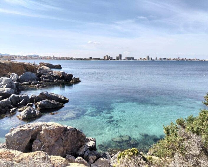 A coastal landscape with clear water, rocky shore, and distant city skyline. 1