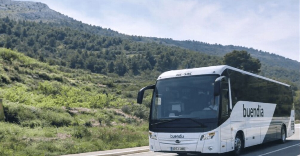A white bus with 'buendia' branding on a road with a hilly landscape in the background. 3