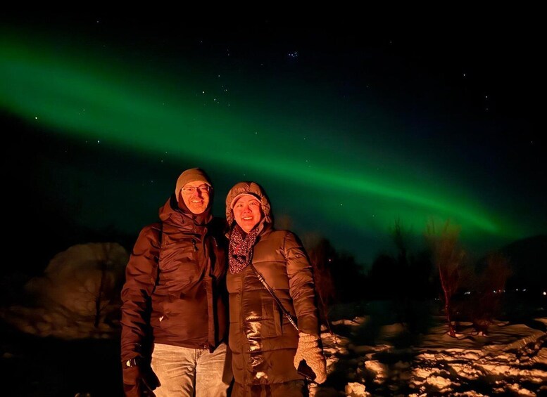Two people standing on snow-covered ground under a night sky with the aurora borealis. 1