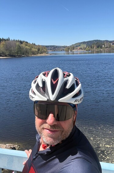 A person wearing a white cycling helmet with 'RUDY' on it, standing by a lake with a clear sky. 2