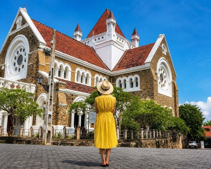 A woman in a yellow dress and hat walking towards a historic building with a red roof and white walls. 4