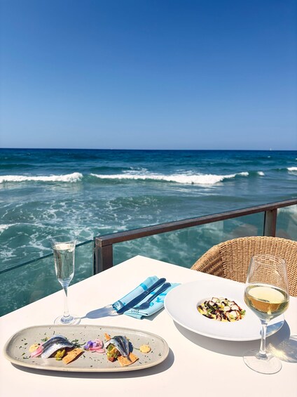 A table with a glass of white wine, a plate of food, and a wicker chair overlooking the ocean. 1