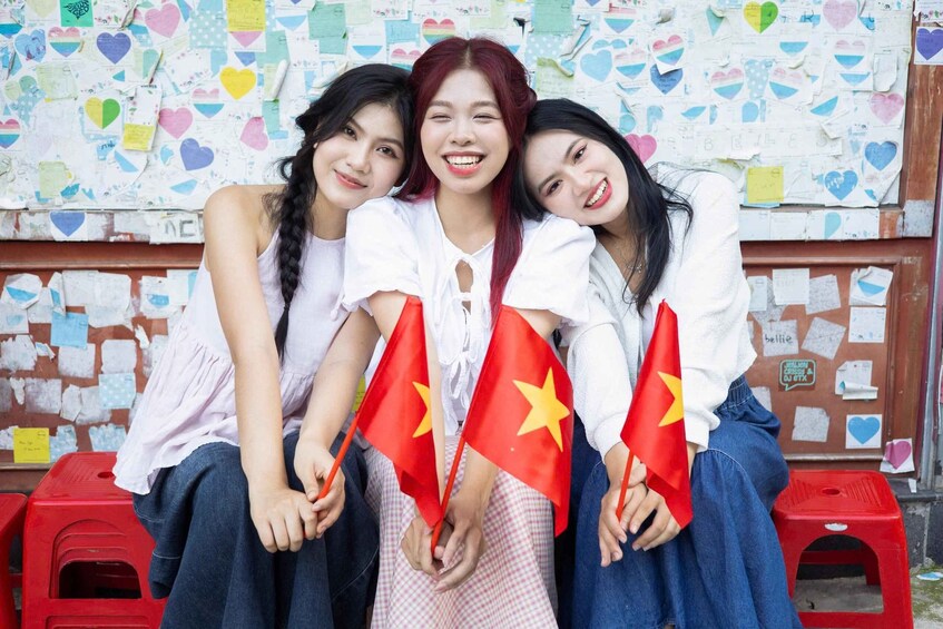 Three individuals sitting on red stools, holding Vietnamese flags, with a wall covered in colorful heart-shaped notes in the background. 5